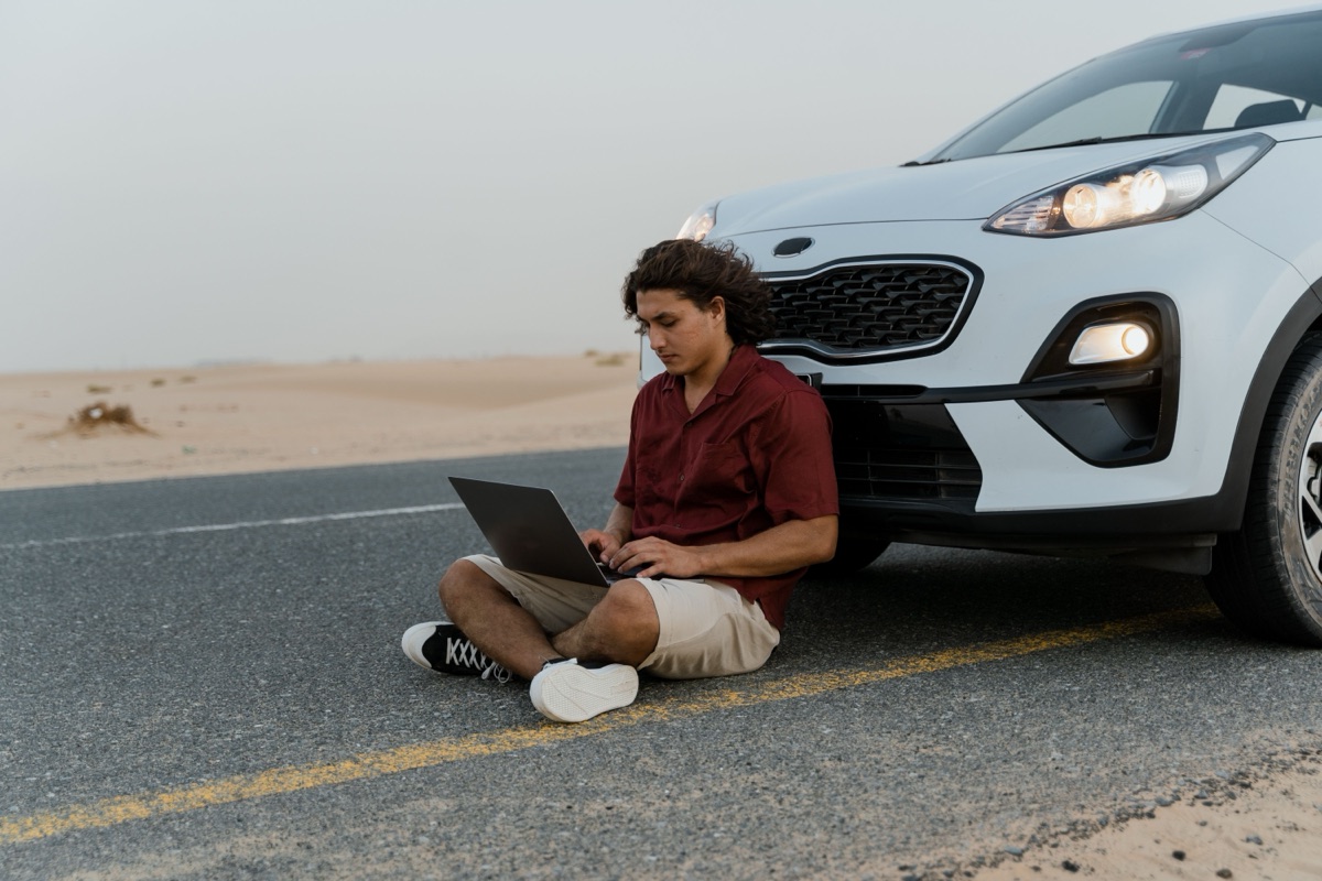 A person working on a laptop while leaning against a car on an open desert road