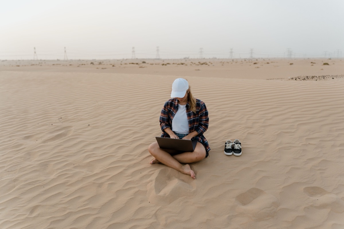 A person sitting cross-legged on a desert dune working on a laptop at dusk