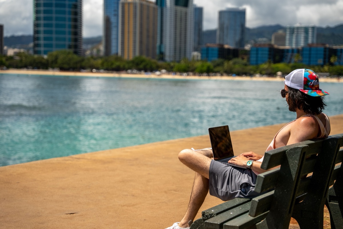 A person working on a laptop on a beachfront bench with a city skyline across the water